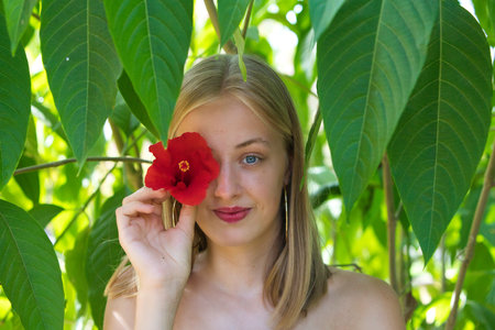 Portrait of a young, beautiful blonde woman with makeup and blue eyes, covering one eye with a red flower. The woman is surrounded by lush vegetation in the park and is smilingの写真素材