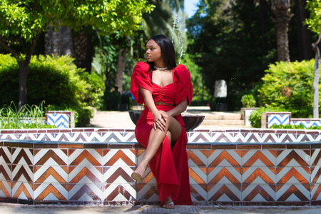 African woman, with long brown hair, young and beautiful dressed in an elegant red dress sitting cross-legged on the edge of a golden tiled fountain in Seville.の写真素材