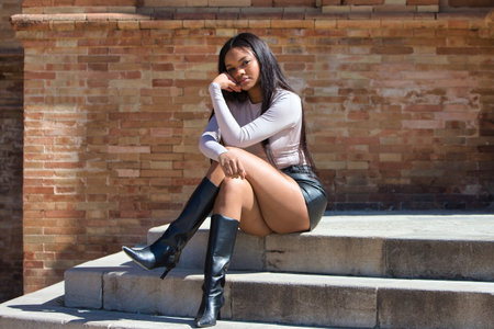 young, beautiful African woman in modern clothes, sitting cross-legged on a staircase in the square of Spain in Seville, red brick wall in the background. Travel and holiday conceptの写真素材