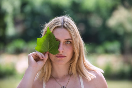 Portrait of a woman covering her face with a leaf. The woman looks serious and hides herself while looking at the camera. In the background is an old, crumbling white wallの写真素材