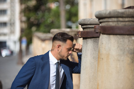 A young, handsome Latin man wearing a suit and tie rests his head on his fist on a stone column. The man looks serious and angry as he stares downwardsの写真素材