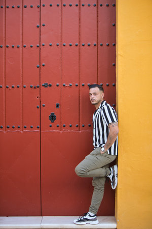 A young, handsome man with a beard, wearing a striped shirt and cargo trousers, rests with one leg on the doorframe of a house with a red Mediterranean-style door in Seville, Spainの写真素材