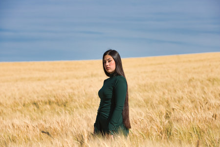 A beautiful, dark-haired teenage Latin girl dressed in green stands among a dry, yellow wheat field on a summer day. In the background, the blue skyの写真素材