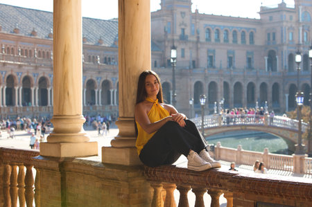 A young, dark-haired, attractive Latin woman wearing a yellow shirt and black trousers is sitting on the railing of the Plaza de España in Seville with her back leaning against a marble columnの写真素材