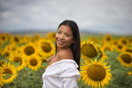 A beautiful young Latin woman with dark skin, wearing a white shirt and bare shoulders, stands among sunflowers on a sunny day. The woman smiles and looks happyの写真素材