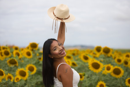 A beautiful black Latin woman wearing a white top and a straw hat stands among sunflowers. The woman is happy and smiling, looking back and lifting her hat with one handの写真素材