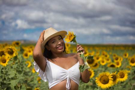 Portrait of a young, dark-skinned Latin woman farmer dressed in a white shirt and straw hat standing among sunflowers. The girl is holding a flower in her hand and is smelling and looking at itの写真素材