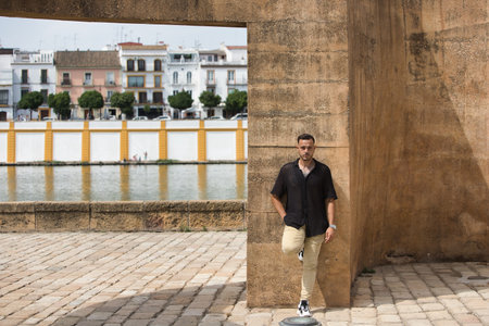 A young, handsome man with a beard, tattoos, earrings and modern clothing is leaning on a brown concrete street structure next to the river. The man is looking in different directionsの写真素材
