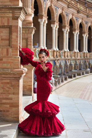 A beautiful woman dancing flamenco leaning against a brick column in a square in Seville, Spain. She is wearing a red ruffled dress.の写真素材