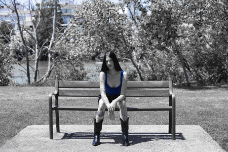 Young, attractive Spanish woman with dark hair, tattoos, wearing a blue top, shorts and black boots, sitting on a park bench sunbathing. In the background, the river and vegetation. Monochrome photoの写真素材