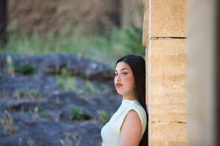 A beautiful Latin teenager with long dark hair leans her back against the stone wall of a ruined castle outside the city. The woman looks at the camera.の写真素材