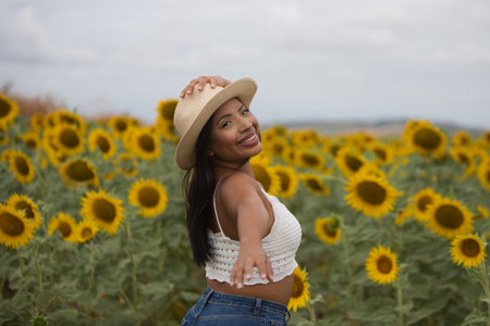 A beautiful black woman wearing a white top and a straw hat stands among sunflowers. The woman is happy and smiling, looking back and holding her hat with one handの写真素材