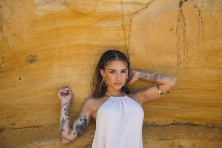 A beautiful young woman wearing makeup, a beige dress and gold jewelry rests against a cliff wall by the sea. The woman strikes different poses and expressions. Concept beauty and fashionの写真素材