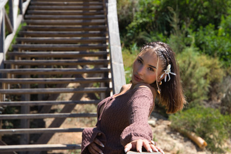 A beautiful young woman with a flower in her hair, wearing a brown dress and a gold belt, rests leaning against a wooden ladder among the vegetation. The woman strikes different poses and expressionsの写真素材