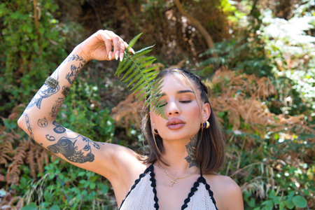 A beautiful young woman covers half her face with a green fern leaf. The woman is happy and has her eyes closedの写真素材