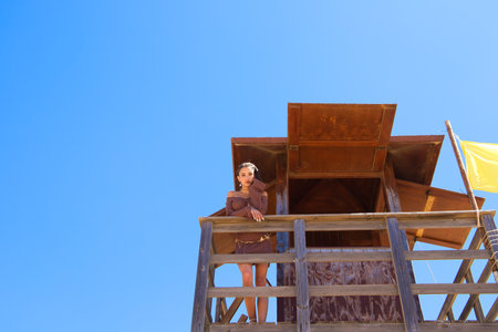 A beautiful young woman in a brown dress looks out to sea from the wooden lifeguard tower with a yellow flag. In the background is a blue skyの写真素材