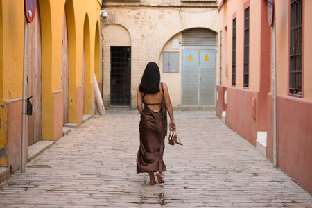 A young, dark-haired woman wearing a brown dress and holding gold heels in her hand, with her back turned, walks barefoot down a lonely alleyway. Her feet plant firmly on the groundの写真素材