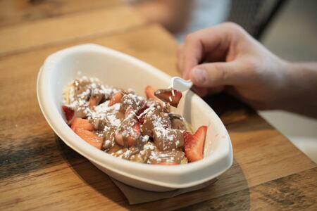 Person eating poffertjes with a plastic fork, poffertjes have chocolate, strawberries and powdered...