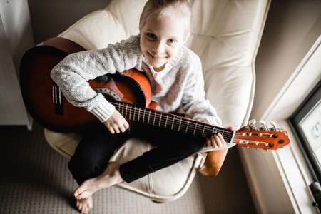 A young blonde caucasion girl sitting by a window practicing her acoustic guitarの写真素材