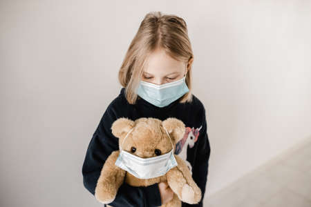 Young caucasion girl wearing a mask and holding a teddy bear who is also wearing a surgical mask during the COVID-19 corona virus pandemicの写真素材