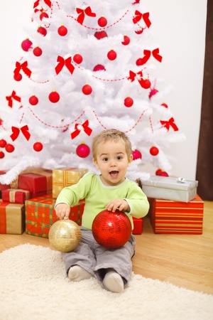 Happy little boy sitting in front of Christmas tree, holding big red and golden ornamentsの写真素材