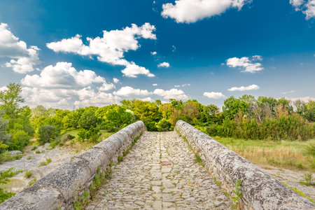 Rock stone bridge across small pond with green trees under blue sky. Historic scenery, idyllic