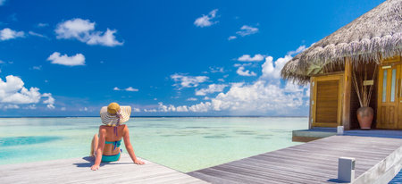 Summer travel vacation. Woman looking at sea view exotic destination. Attractive woman sitting enjoying sunlight lifestyle wearing bikini hat. Leisure beach seascape water villaの写真素材