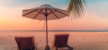 Sunset beach. Beautiful tropical island two sun beds lounger, parasol under palm tree. Sand sea colorful dream sky, calm relax. Summer vacation landscape. Romantic couple resortの写真素材