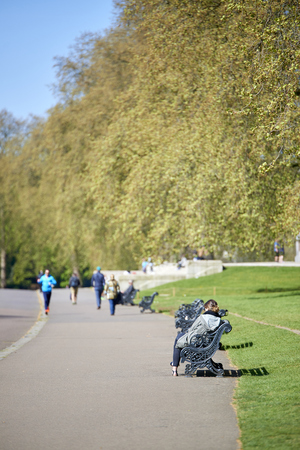 LONDON, UK - APRIL 22: Kensington Park visitor sitting on bench on sunny spring day, with people walking in the pavement in the tree lined blurry background. April 22, 2015 in London.のeditorial素材