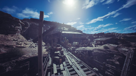 A deserted mining town features wooden structures and scattered supplies. The bright sun shines above a dusty path leading through rugged terrain and natural rock formations.の写真素材