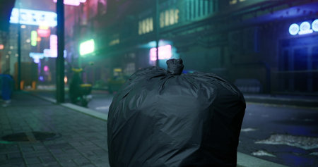 In a dimly lit urban environment, a large black trash bag sits on the sidewalk. Neon signs glow in the background, reflecting an almost eerie atmosphere during late night hours.の写真素材