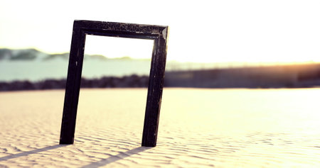 A striking black frame stands alone in a vast desert landscape, silhouetted against a stunning sunset. Soft sand ripples create patterns extending towards distant hills, evoking tranquility.の写真素材