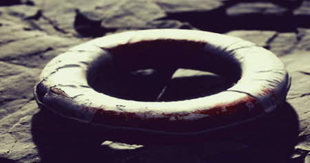 A worn lifebuoy rests on sunlit, jagged rocks beside a tranquil body of water as the sun sets, casting a warm glow and creating reflections on the surface.の写真素材