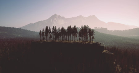Vibrant trees stand proudly atop a rugged cliff, framed by majestic mountains under a pastel sky. The serene landscape captures the essence of natures beauty at dusk.の写真素材