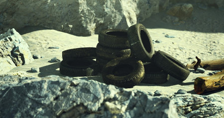 A pile of worn out tires rests on a sandy beach surrounded by rocks. Sunlight casts shadows,...