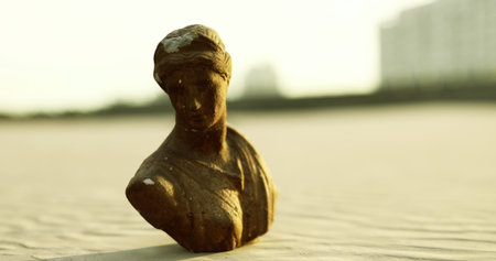 A weathered bronze bust of a woman stands prominently amidst the golden sand, illuminated by soft rays of the setting sun. Gentle waves lap at the shore nearby.の写真素材