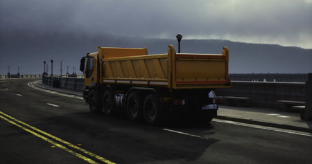 A vibrant yellow dump truck navigates a deserted coastal road. The gloomy clouds hang low in the sky, creating an atmospheric backdrop. Street lamps line the roadway, adding to the serene ambiance.の写真素材