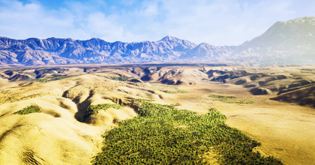 Expansive desert terrain unfolds under a bright blue sky, with verdant patches of vegetation contrasting against rugged mountains in the distance, creating a serene and captivating vista.の写真素材