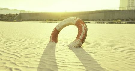 A weathered lifebuoy stands alone in soft golden sands as the sun sets on the horizon. Long shadows stretch across the beach, creating a tranquil atmosphere filled with reflection.の写真素材