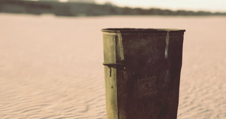 A rusted trash can, weathered and worn, stands solitary on a vast expanse of soft sand at dusk, with golden light casting long shadows. Nature slowly reclaims its space and beauty.の写真素材