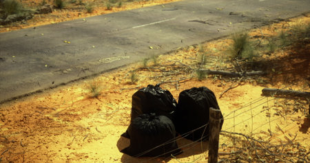 In a remote desert setting, three bags filled with trash sit abandoned next to an empty road. Sunlight casts long shadows and highlights the contrasting colors of the dry terrain.の写真素材
