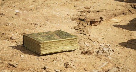 An aged book rests on dry, cracked earth surrounded by rocky terrain. Sunlight illuminates its...