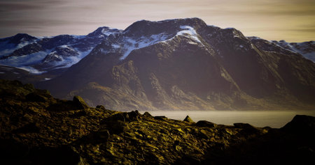 Reflective waters mirror the rugged mountain peak bathed in warm golden light. The rocky foreground adds depth, while snow caps hint at the chilling beauty of Norways nature.の写真素材