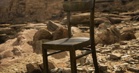 A wooden chair stands alone amidst a rugged landscape filled with stones and rocks. The bright sunlight highlights the chairs features against the barren terrain, creating a striking contrast.の写真素材