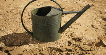 A weathered metal watering can lies on cracked, dry earth bathed in warm sunlight. The scene reflects the challenges of gardening in a dry environment, emphasizing resilience.の写真素材