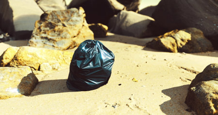 A black trash bag sits alone on the sandy beach, surrounded by various rocks. The bright sunlight illuminates the scene, emphasizing the contrast between the bag and natural elements.の写真素材