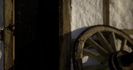 A weathered wooden wheel rests beside a rustic door, casting shadows on the textured wall. The warm light of sunset enhances the villages historic charm and tranquility.の写真素材