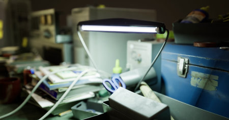 In a warmly lit workspace, a bright desk lamp casts light on various tools and materials scattered across the desk. Papers, scissors, and a toolbox indicate ongoing projects.の写真素材
