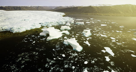 Large icebergs float in near freezing waters as the sun sets behind distant mountains, creating a...