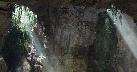 Bright rays of sunlight pour into a rocky cave, illuminating the damp stones and lush greenery...
